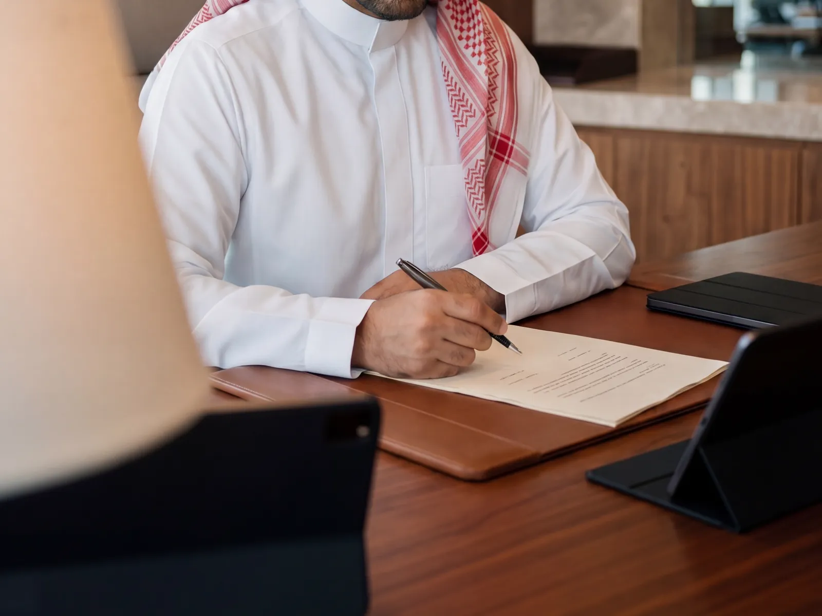 محامٍ يرتدي الزي السعودي يدوّن ملاحظاته بالقلم على مستند قانوني فوق مكتب خشبي داخل مكتب المحاماة